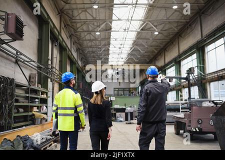 Rear view of manager supervisors and industrial worker in uniform walking in large metal factory hall and talking. Stock Photo