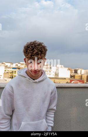 Portrait of smiling young Caucasian man with curly hair dressed in a white sweatshirt looking at the camera with the city skyline in the background. Stock Photo
