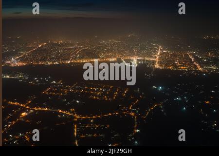 Aerial view from airplane window of buildings and bright illuminated streets in city residential area at night. Dark urban landscape at high altitude. Stock Photo