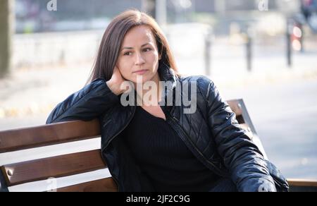Young sad woman sitting alone on street bench outdoors thinking about something. Loneliness and depression during hard period in life concept Stock Photo