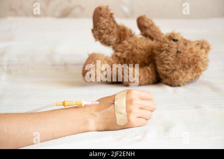 Children's hand with a dropper on the bed and next to it lies a children's teddy bear in the hospital in the ward, children's medicine, a sick child Stock Photo