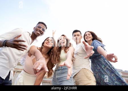 Low angle view of a group of multiracial friends looking at camera and smiling while enjoying time together outdoors. Friendship concept. Stock Photo