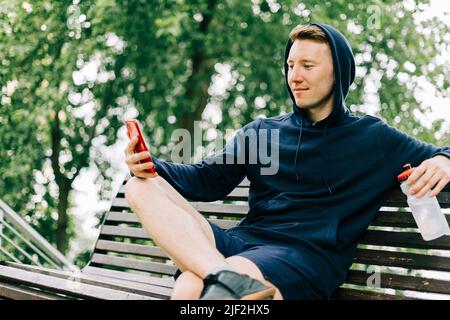 Happy Young man runner in hoodie sitting on bench and relaxing after sport training, using mobile phone. Holding water bottle while doing workout in Stock Photo