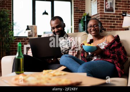 Man working on laptop and woman switching tv channel program, eating takeout delivery meal and watching movie on television. Enjoying fast food and beer, using computer at home. Stock Photo