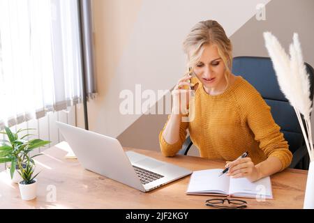 Attractive european business woman is on phone and taking notes at same time writing down important information. Modern electronic gadgets and communication in a modern office in a yellow jacket Stock Photo