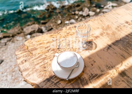 Cappuccino latte coffee cup mug and a glass of water standing on a wooden table in outside cafe with scenery sea and mountains in the background. Copy Stock Photo