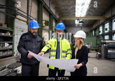 Manager supervisor, engineer and industrial worker in uniform discussing blueprints in large metal factory hall. Stock Photo
