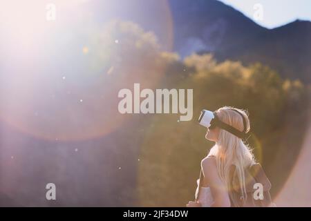 Blonde woman wearing VR headset during a backpacking holiday. Woman hiking through mountains wearing VR headset. Woman on holiday using virtual Stock Photo