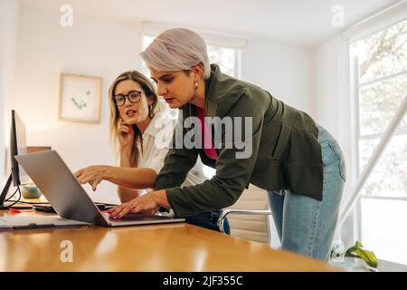 Two young businesswomen collaborating on a new task in a creative office. Female entrepreneurs using a laptop while working as a team in a woman-owned Stock Photo
