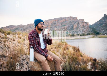 A young adult man takes a break from hiking drinks coffee by the lake view Stock Photo