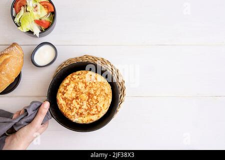 Women hand holding frying pan with Tortilla, Spanish omelette made with eggs and potatoes and served with  salad and bread. Top view. Stock Photo