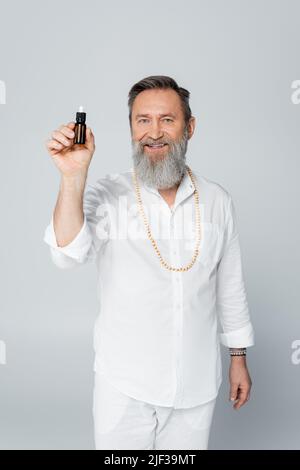 bearded healing guru holding bottle of essential oil and smiling at camera isolated on grey Stock Photo