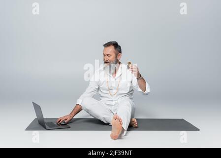 barefoot healing guru sitting near laptop and holding bottle of scented oil on grey Stock Photo