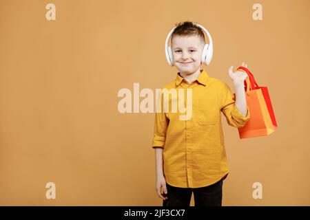 Image of smiling boy holding bags with presents or shoppings on yellow background. child in white headphone. Shopper with many colored paper bags. Holidays sales and discounts. Cyber monday. Stock Photo