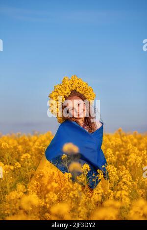 Ukrainian child girl wrapped in blue and yellow Ukrainian flag in yellow wreath in field of yellow flowers against blue sky. Pray for Ukraine. Ukraine's Independence Flag Day. Symbols of Ukraine. Stock Photo