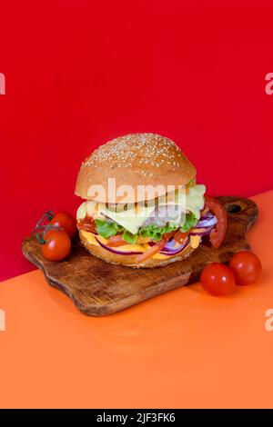 Delicious burger on the board with cutlet, tomatoes and cheese, cucumber and lettuce. On a red background. Fast food. copy space Stock Photo