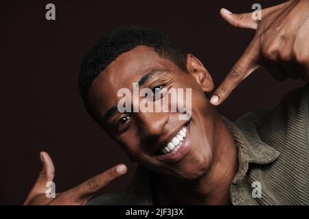 Young black man dressed in shirt pointing fingers at his smile isolated over brown wall Stock Photo