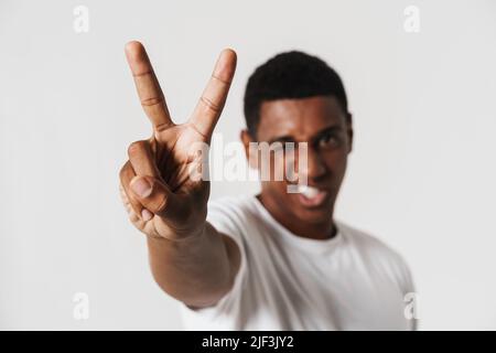 Young black man grimacing while showing peace sign isolated over white wall Stock Photo