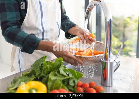 Midsection of african american mid adult man wearing apron cleaning carrots in kitchen sink Stock Photo