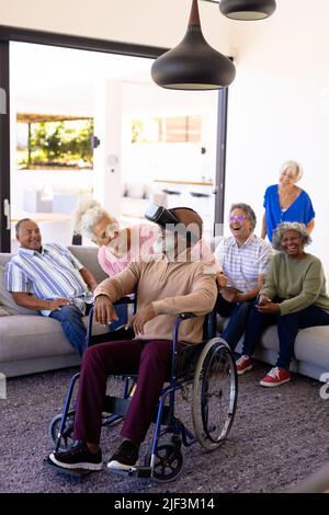 Cheerful multiracial seniors looking at friend using virtual reality while sitting on wheelchair Stock Photo