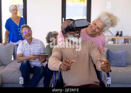 Multiracial seniors looking at friend using virtual reality simulator while sitting on wheelchair Stock Photo