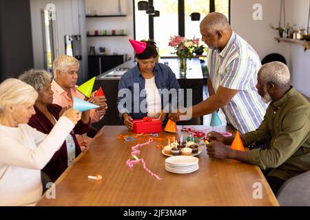Multiracial seniors wearing party hats while woman opening birthday gift at table in nursing home Stock Photo