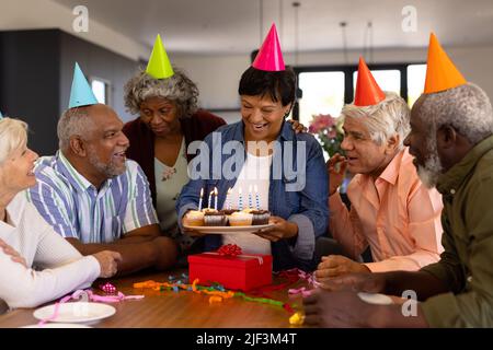 Multiracial seniors wearing hats looking at happy woman holding cupcakes with birthday candles Stock Photo