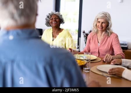 Happy multiracial senior friends talking while sitting at dining table in nursing home Stock Photo