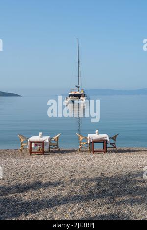 Bodrum, Mugla, Turkey. April 22nd 2022 Restaurant tables on the beach with a Catamaran moored in Bodrum harbor, a sailing and cruise destination Stock Photo
