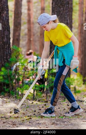 Little girl picks up trash in woods on summer day. Ecological action. Child in yellow T-shirt cleanses nature of pollution and trash. Real scene. Background. Stock Photo