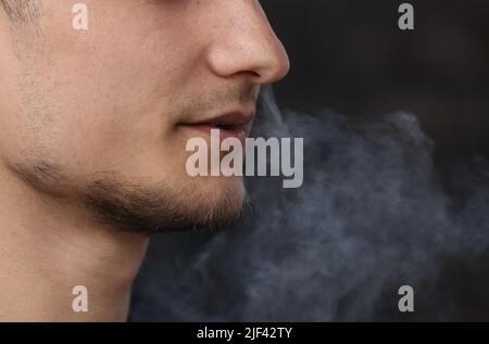 close up of a man with an electronic cigarette. man smokes an electronic cigarette in a natural background. hipster smokes an e-cigarette, breathes out streams of smoke and vaping outdoors copy space. Stock Photo