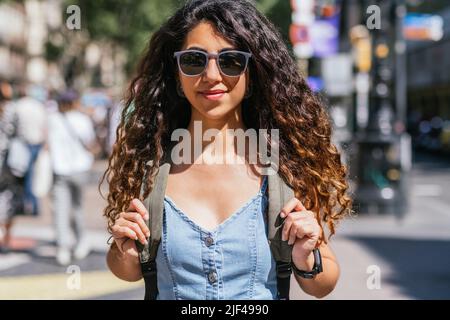 portrait of a beautiful dark haired young woman wearing denim dress. She wears sunglasses and a backpack and looks to camera positively Stock Photo