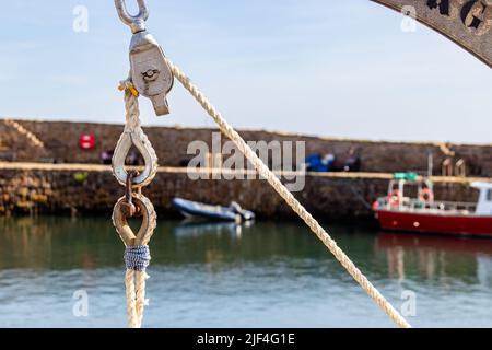 Close up of a rope, pulley and shackle in the sun, against an out of focus harbour wall or quayside with boats in the background, including a dinghy Stock Photo