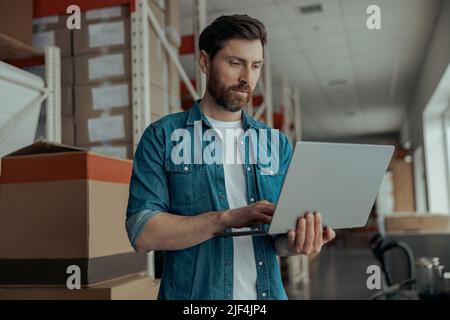Serious man worker working on laptop stands on warehouse  Stock Photo