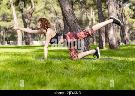 Front view of young woman kneeling on grass stretching leg in yoga  position, looking away, Philadelphia, Pennsylvania, USA stock photo