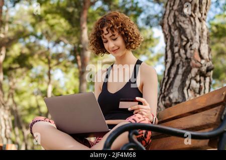 Young beautiful woman wearing sportive clothes on city park, outdoors using laptop computer and holding credit card. Online shopping, banking and tran Stock Photo