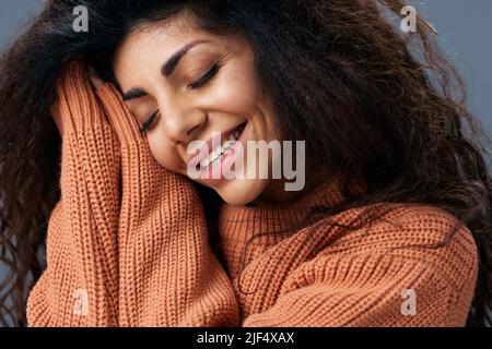 SLEEPY CONCEPT. Cheerful smiling tanned curly Latin female in warm sweater fold hands near cheek posing isolated over gray blue background close eyes Stock Photo