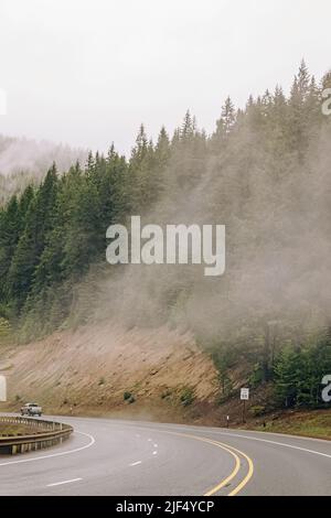 Whining mountain road with a car in a distance. Road trip through the forest of pine trees on a rainy, cloudy and foggy day. Stock Photo