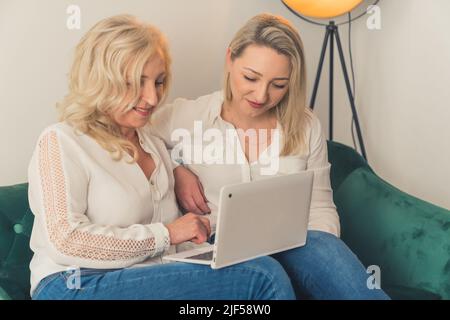 Caucasian beautiful short-haired middle-aged woman sitting on a green couch with a laptop on her legs, explaining something to the other older woman. High quality photo Stock Photo