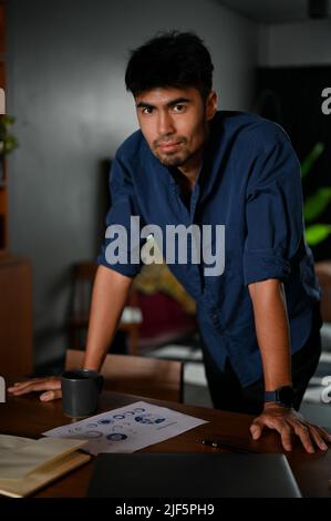 Smart and confident Asian male entrepreneur or businessman in casual standing leaning on the office desk. Stock Photo