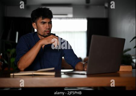 Stressed serious and professional Asian businessman or male tech engineer remote working at his home, using a laptop computer. Stock Photo