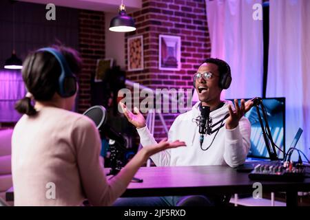 Podcast presenter recording livestream chat at production station, using modern equipment for audience. Male influencer interviewing woman for online broadcast discussion in studio. Stock Photo