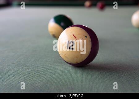 An old billiard ball with the number 12 (twelve) in lilac lies on the faded cloth of the billiard table. Stock Photo