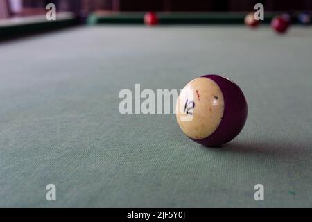 An old billiard ball with the number 12 in lilac lies on the faded cloth of the billiard table. Stock Photo