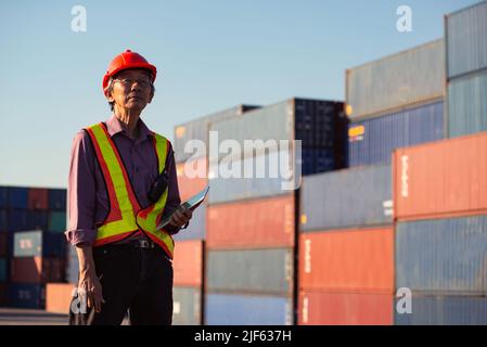 A senior elderly Asian worker engineer wearing safety vest and helmet standing and holding digital tablet at shipping cargo containers yard. Stock Photo