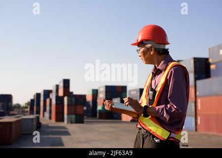 A senior elderly Asian worker engineer wearing safety vest and helmet standing and holding digital tablet at shipping cargo containers yard. Stock Photo