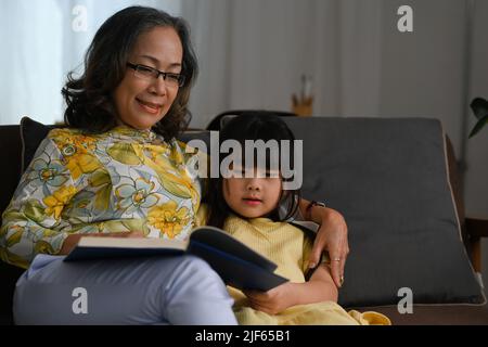 Happy moments with grandmother and little granddaughter reading book together on sofa Stock Photo