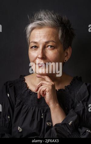 Close up gorgeous mature businesswoman with grey hair posing leaned chin on her hand isolated on black background. Graying elegant woman with short hair. Image of successful mature woman. Aged beauty. Stock Photo