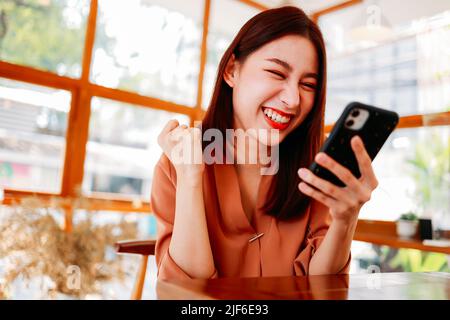 Portrait of cheerful young Asian woman feeling excited on winning gift hamper in smartphone while relaxing in modern cafe Stock Photo