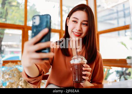 Young Asian female food blogger taking selfie with smartphone holding fresh glass of cocoa while sitting in cafe with social distancing during coronavirus Stock Photo